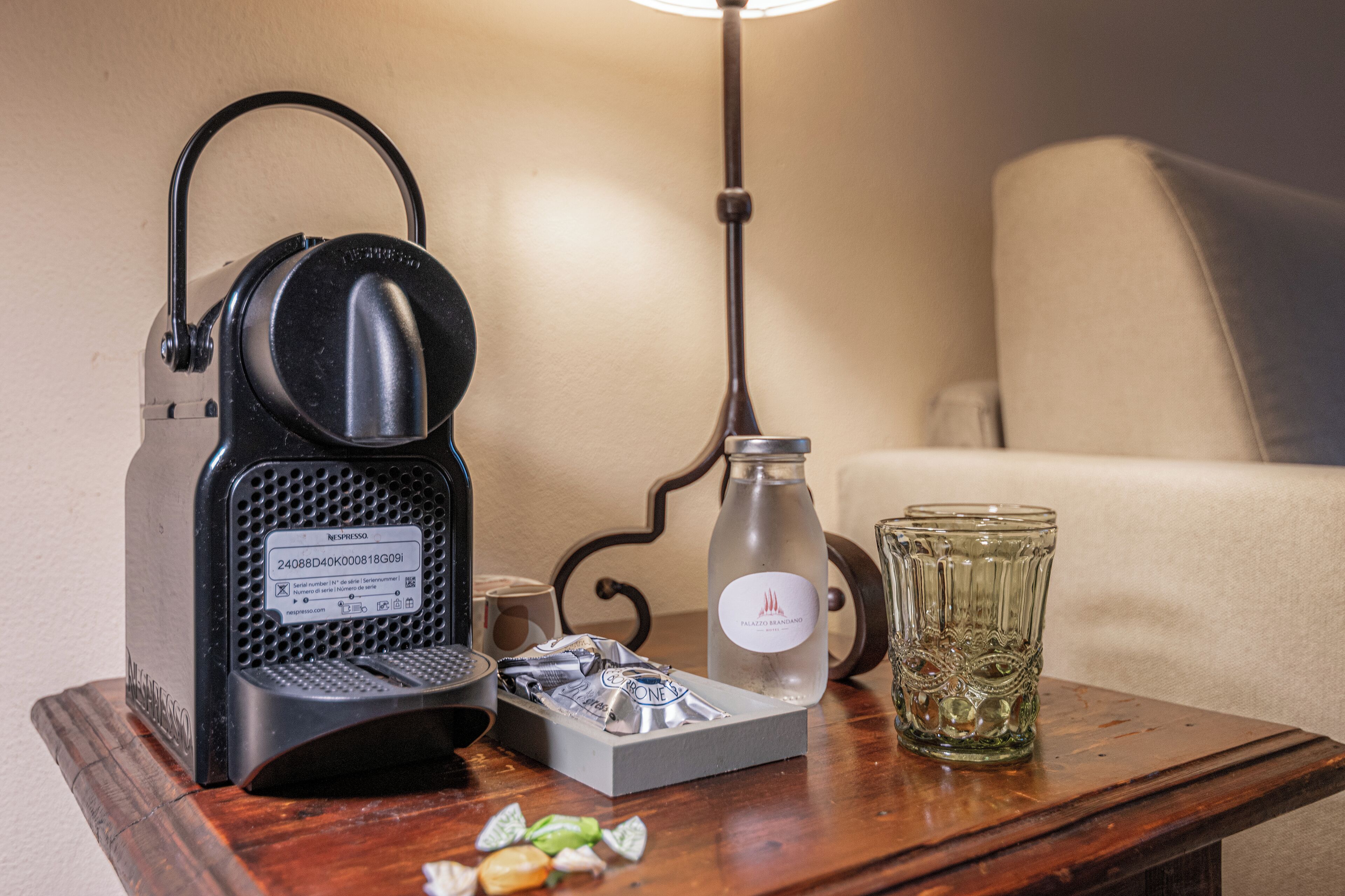 A coffee maker, a glass, and a bottle on a wooden table in a room with a lamp and a chair.