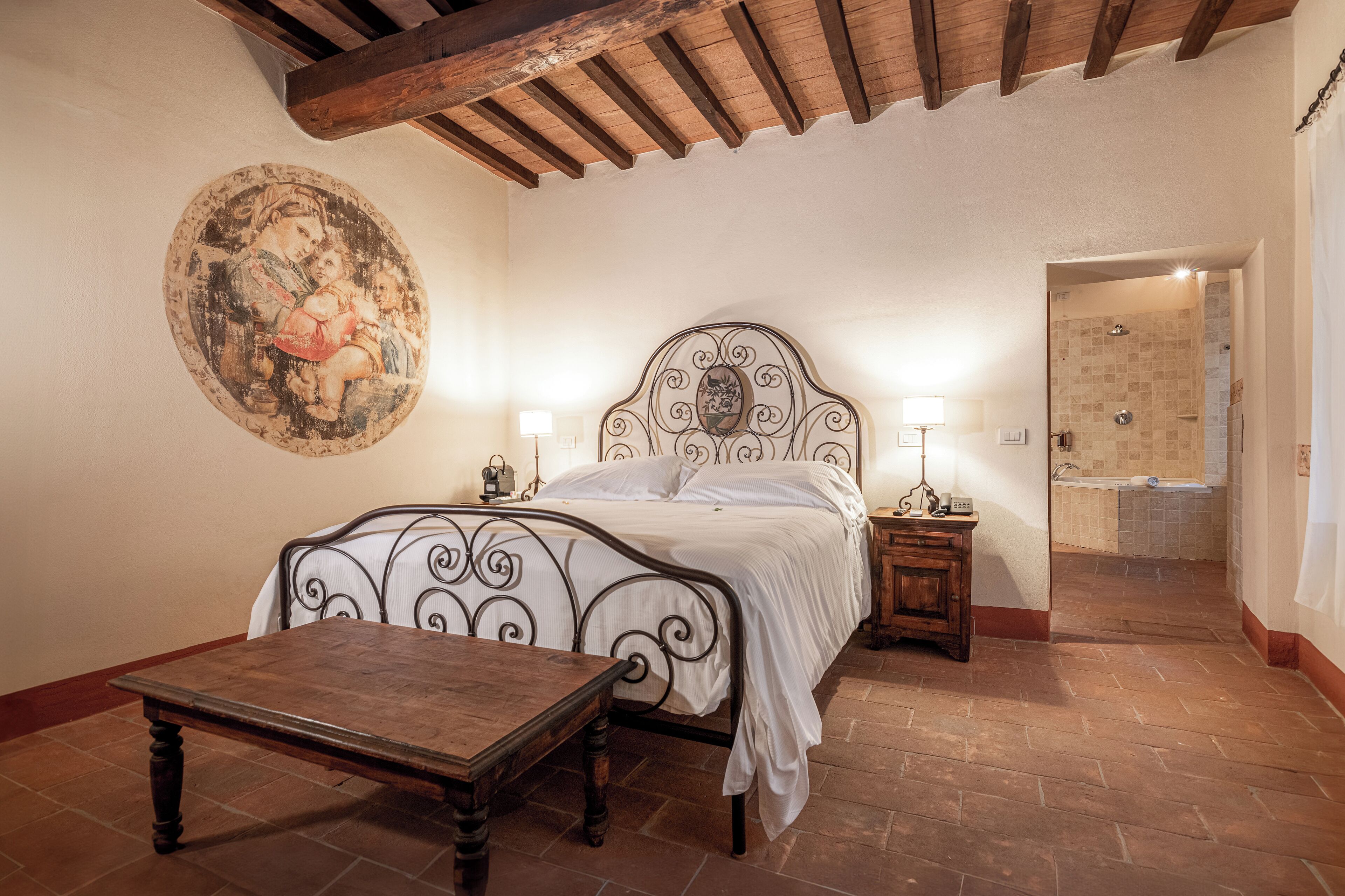 A bedroom with a bed, a wooden table, a wall-mounted painting, and a bathroom visible through an open door.