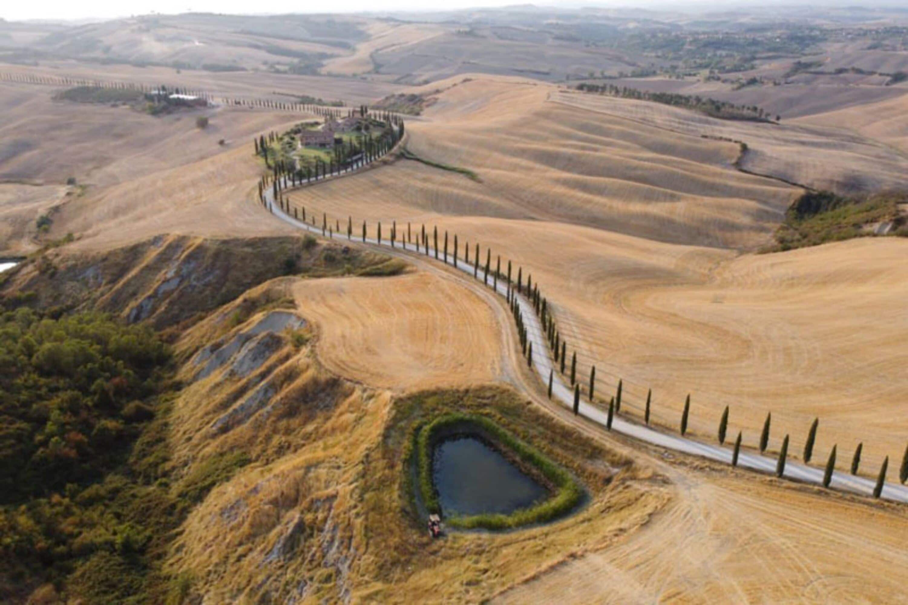 An aerial view of a rural landscape with rolling hills, a winding road, and a fenced area with a small pond.