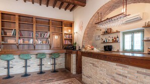 A bar area with wooden shelves, books, and a brick wall. There are three green bar stools and a patterned rug on the floor. - Palazzo Brandano (Trequanda)