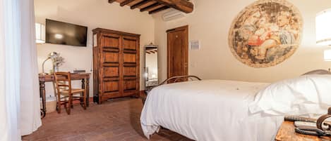 A bedroom with a bed, wooden ceiling beams, a television, a desk with a chair, and a wall-mounted painting.