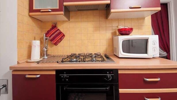 Een keuken met een gasfornuis, een magnetron, een afwasschaal en een rode handdoek aan de muur.