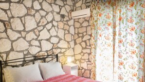 Ein Bett mit gemustertem Überwurf, eine Steinwand, eine Klimaanlage, ein Nachttisch mit Lampe und blumige Vorhänge.