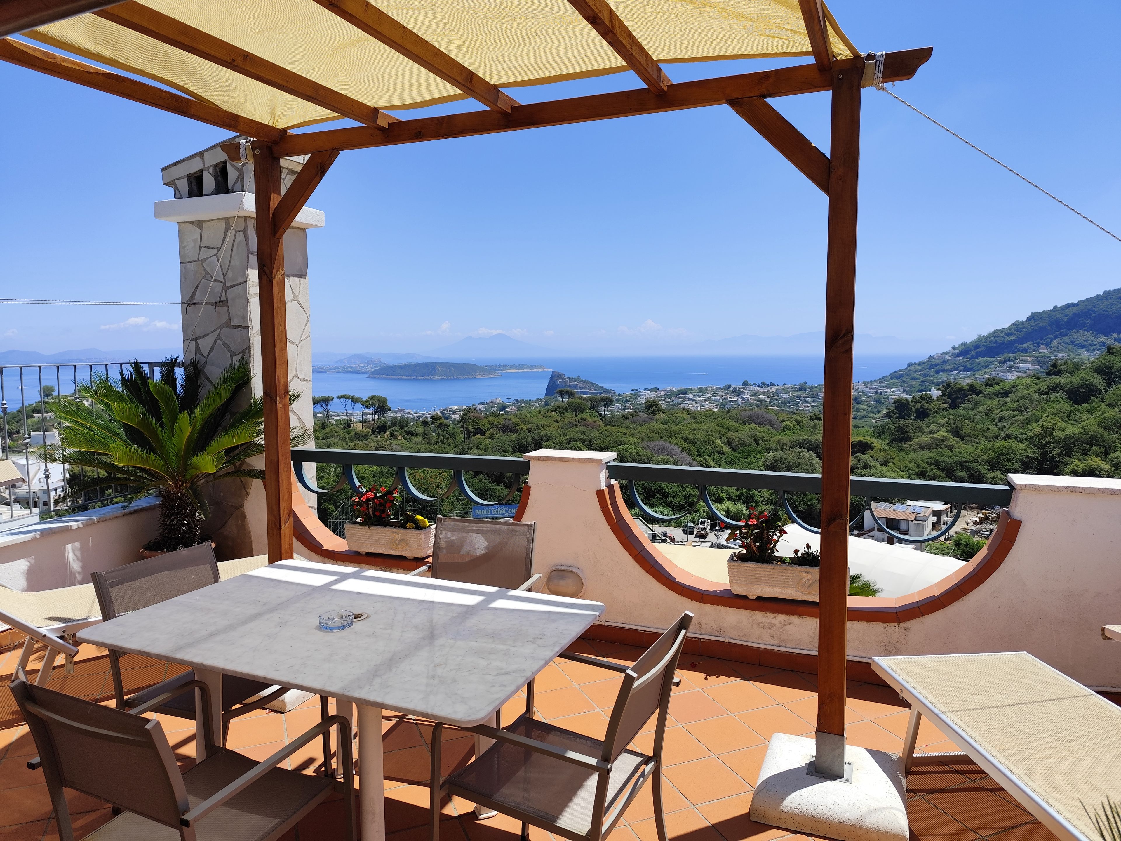 A terrace with a table and chairs, overlooking a scenic view of the sea and hills.