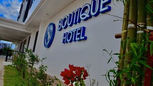 A sign for Boutique Hotel with a logo, surrounded by greenery and a clear sky.