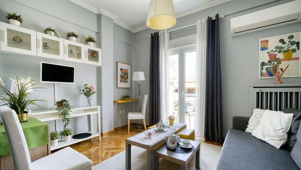 A modern living room with a grey sofa, a dining table, and a TV mounted on the wall. There are potted plants on shelves and a window with curtains. - 1 Bedroom Superior Apartment (Athens)