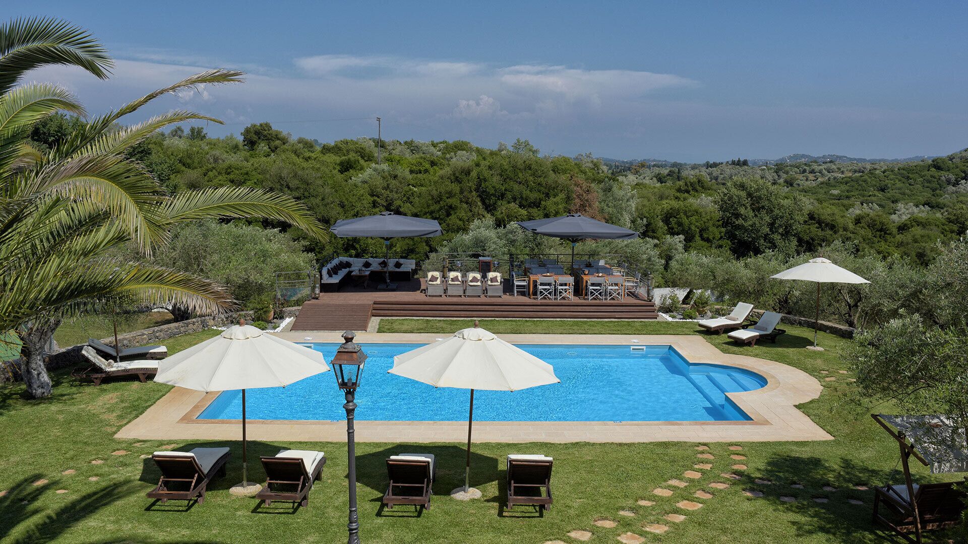 Ein Pool mit Liegestühlen und Sonnenschirmen, umgeben von üppiger Vegetation und Hügeln im Hintergrund.