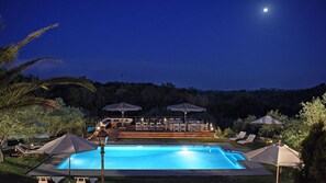 A nighttime pool area with lounge chairs and umbrellas, surrounded by greenery and hills. - Vip Luxury Villa Privilege Classic Exclusive Corfu (Corfu)