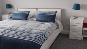 Un dormitorio con una cama, dos mesitas de noche, una lámpara en cada una, suelo alfombrado y dos cuadros en la pared.