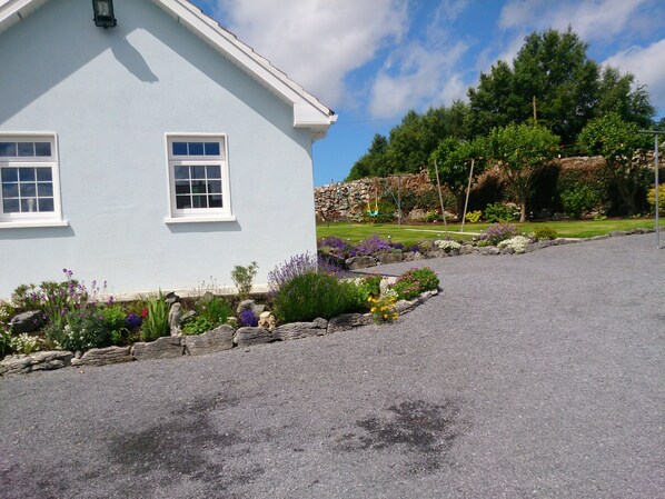 A light blue building with two windows, a stone wall, and a garden with various plants and flowers.