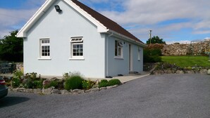 Una casa de un piso con fachada blanca y techo marrón, un camino de entrada y un muro de piedra al fondo.