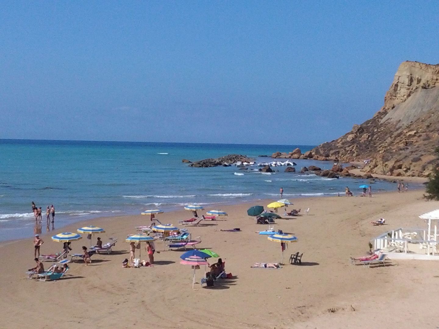 A beach with multiple umbrellas and sun loungers, rocky cliffs in the background, and clear blue skies.