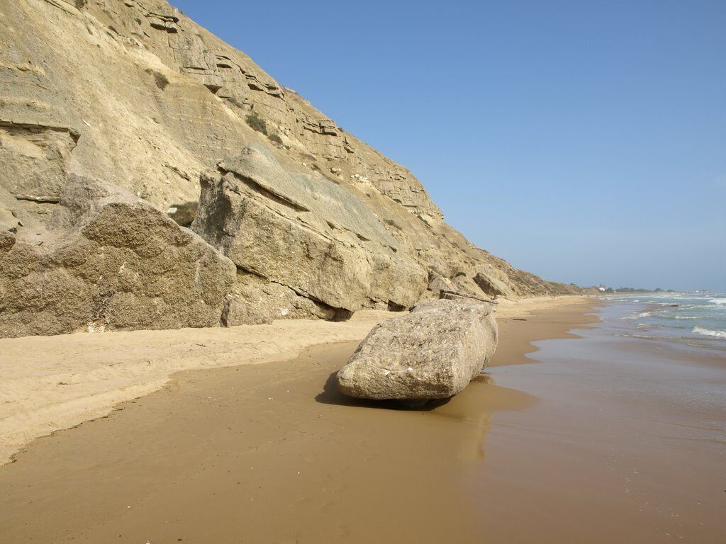 A beach with a large rock in the foreground and a rocky cliff in the background.
