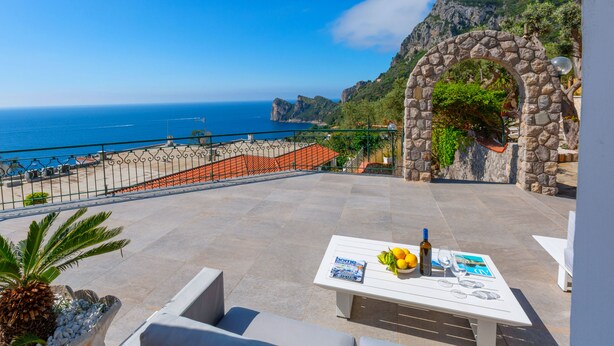 A terrace with a table set for two, overlooking a coastal view with a stone archway and a palm tree. - Villa Marianna Like Paradise in Amalfi Coast!!! (Massa Lubrense)