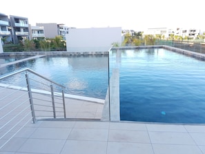 Ein Schwimmbecken mit Metallleiter und Fliesenveranda, umgeben von Gebäuden und Grünflächen.