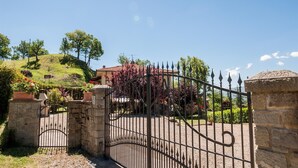 A gated entrance with a stone wall, a metal gate, and a house with a red-tiled roof surrounded by greenery and flowering plants. - Wonderful Villa With Private Pool in the Heart of Tuscany (Pratovecchio Stia)
