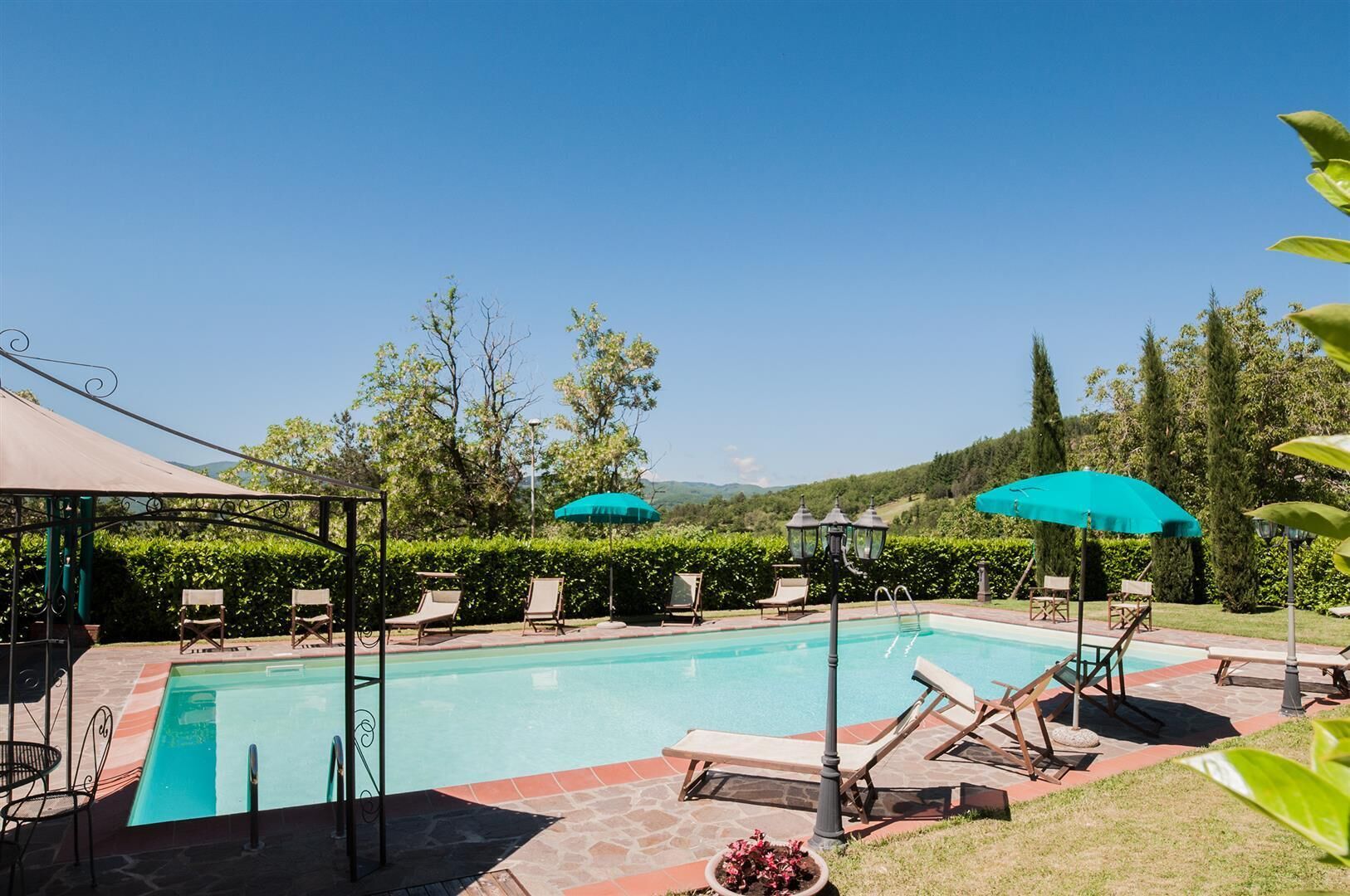 Piscina con sillones de descanso y sombrillas, rodeada de vegetación y un cielo despejado.