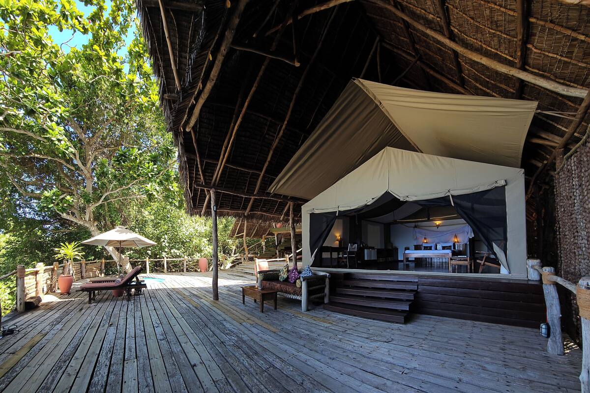A wooden deck with a covered seating area, a large tree, and a thatched roof structure.