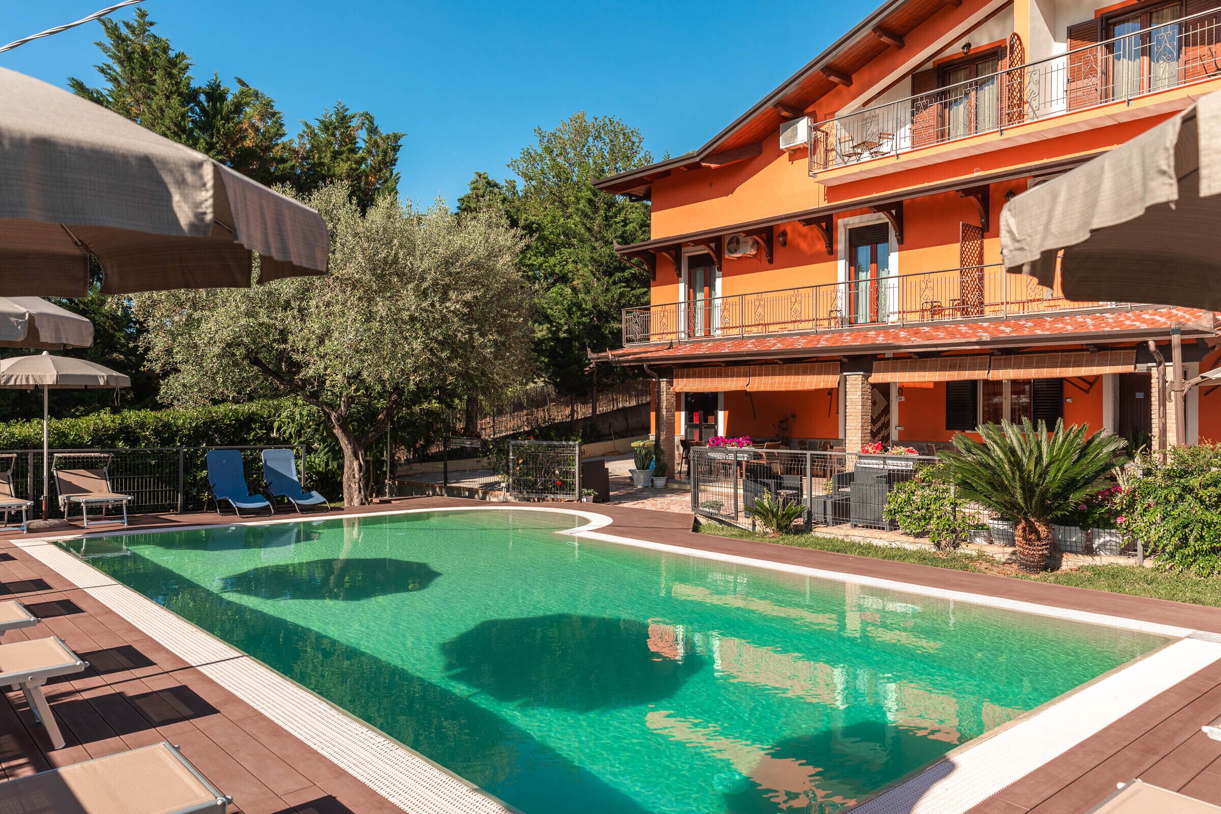 A swimming pool with lounge chairs and umbrellas, surrounded by a building with balconies and a garden.
