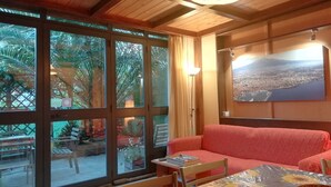 A room with a wooden ceiling, a red sofa, a dining table with chairs, a large window with a view of palm trees, and a framed cityscape picture on the wall. - House Between Etna and the sea Near Taormina (Santa Venerina)