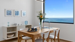Un espace repas moderne avec une table en bois, des chaises blanches et une vue sur l’océan grâce à une grande fenêtre.