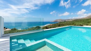 Une piscine à débordement moderne, avec une eau d’un bleu cristallin, entourée d’une balustrade en verre, offrant une vue panoramique sur un paysage côtier magnifique, avec des montagnes et une végétation luxuriante.