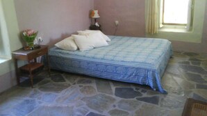 A bedroom with a stone floor, a bed with a blue patterned bedspread, a nightstand with a lamp, and a window with curtains. - Beautiful Country Home on Syros Island, Greece (Syros)