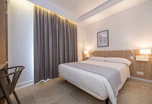 A hotel room with a bed, two bedside lamps, a chair, and a large window with curtains. - Hotel Victori (Es Castell)
