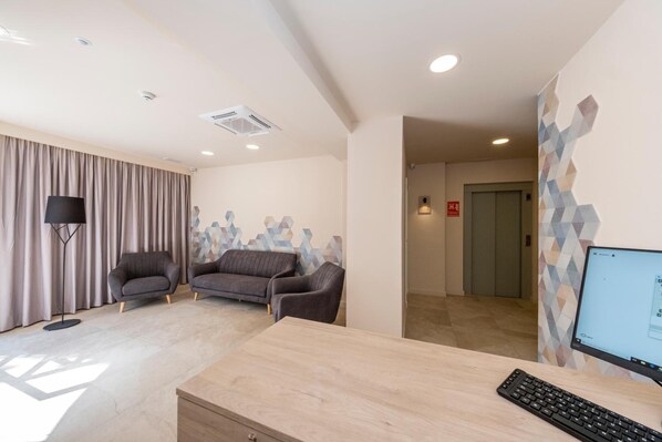 Habitación de hotel moderna con escritorio, dos sillones, un ventanal con cortinas y una pared con diseño geométrico.