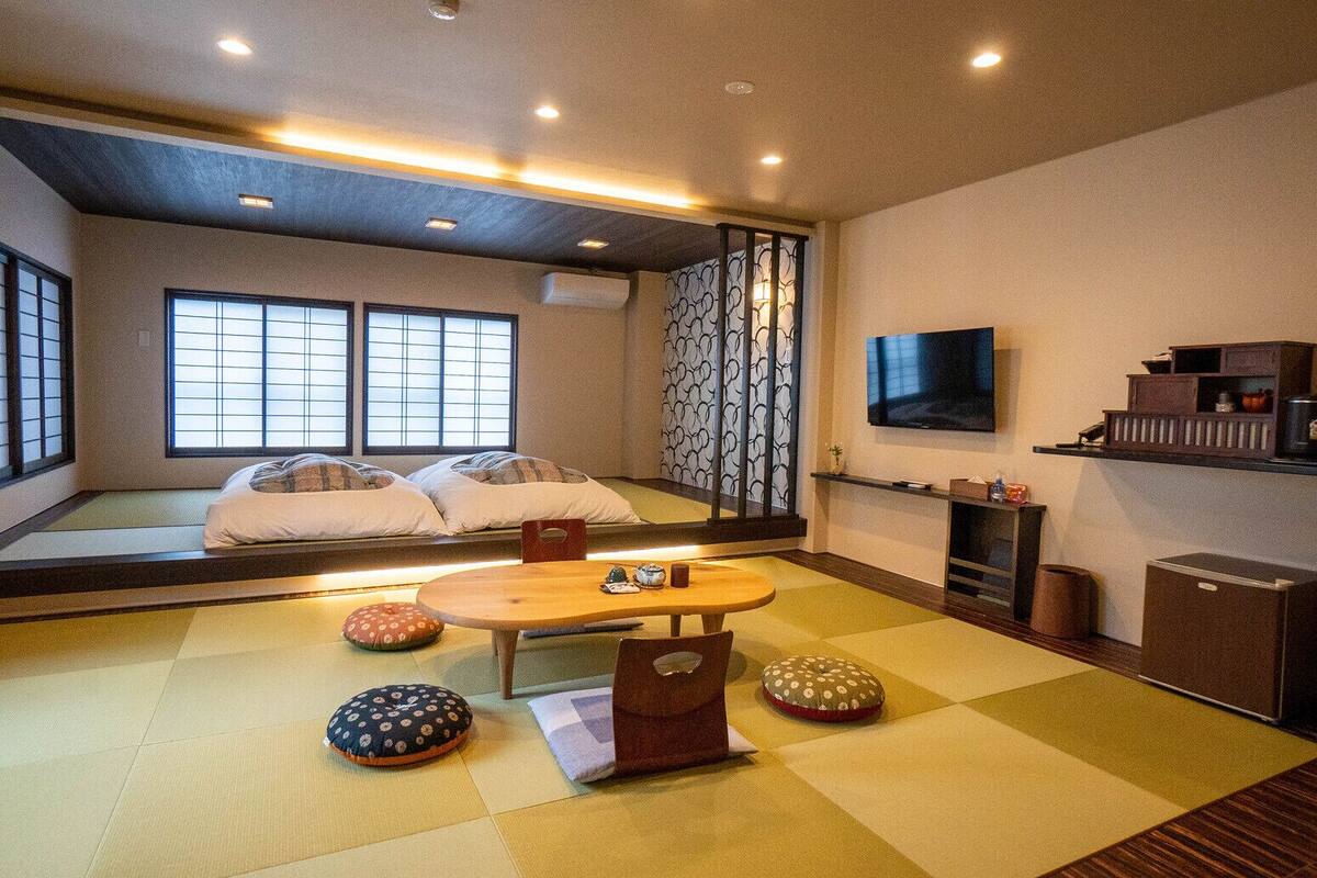 A traditional Japanese room with tatami flooring, a low table, cushions, a TV, and a sliding door.