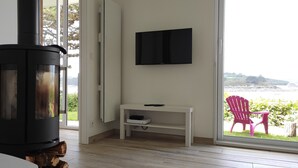 Una sala de estar moderna con una estufa de leña, un televisor de pantalla plana montado en la pared y vistas a un área verde con una silla afuera.