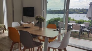 Habitación moderna con una mesa de comedor de madera, sillas blancas y anaranjadas, un televisor en la pared y vista a barcos en un lago.