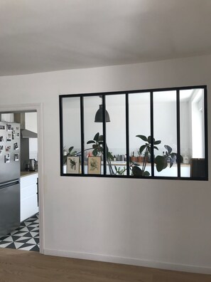 Modern kitchen with a large window offering a view of the adjacent living space, featuring a stainless steel refrigerator, coffee machine, and potted plants.