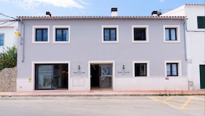 Un edificio de dos pisos con techo plano y fachada de tejas. Hay cuatro ventanas en el piso superior y tres en la planta baja. La entrada tiene una puerta de vidrio y un letrero que dice "HOTEL SON VILAR".