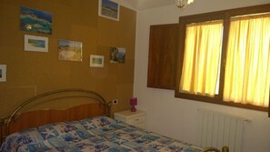 A bedroom with a bed, a nightstand, a lamp, a window with curtains, and wall-mounted pictures. - Portion of a Villa Near the sea (Capoterra)