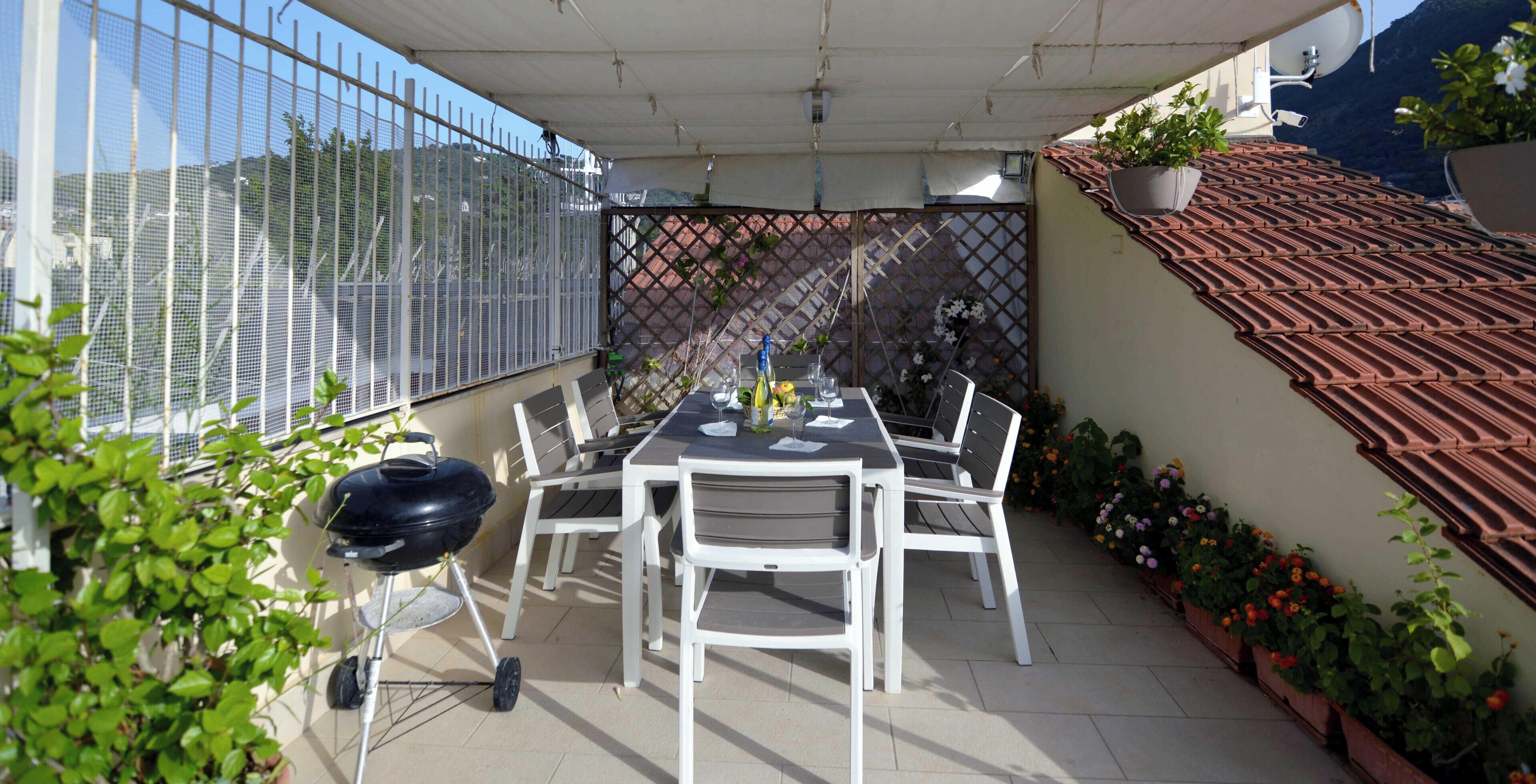 A covered patio with a table and chairs, a grill, and potted plants.