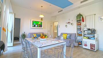 A modern living room with a glass dining table, clear chairs, a sofa, and a bookshelf.