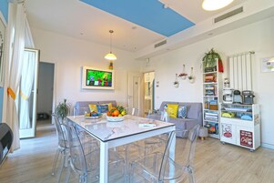 A modern living room with a glass dining table, clear chairs, a sofa, and a bookshelf.
