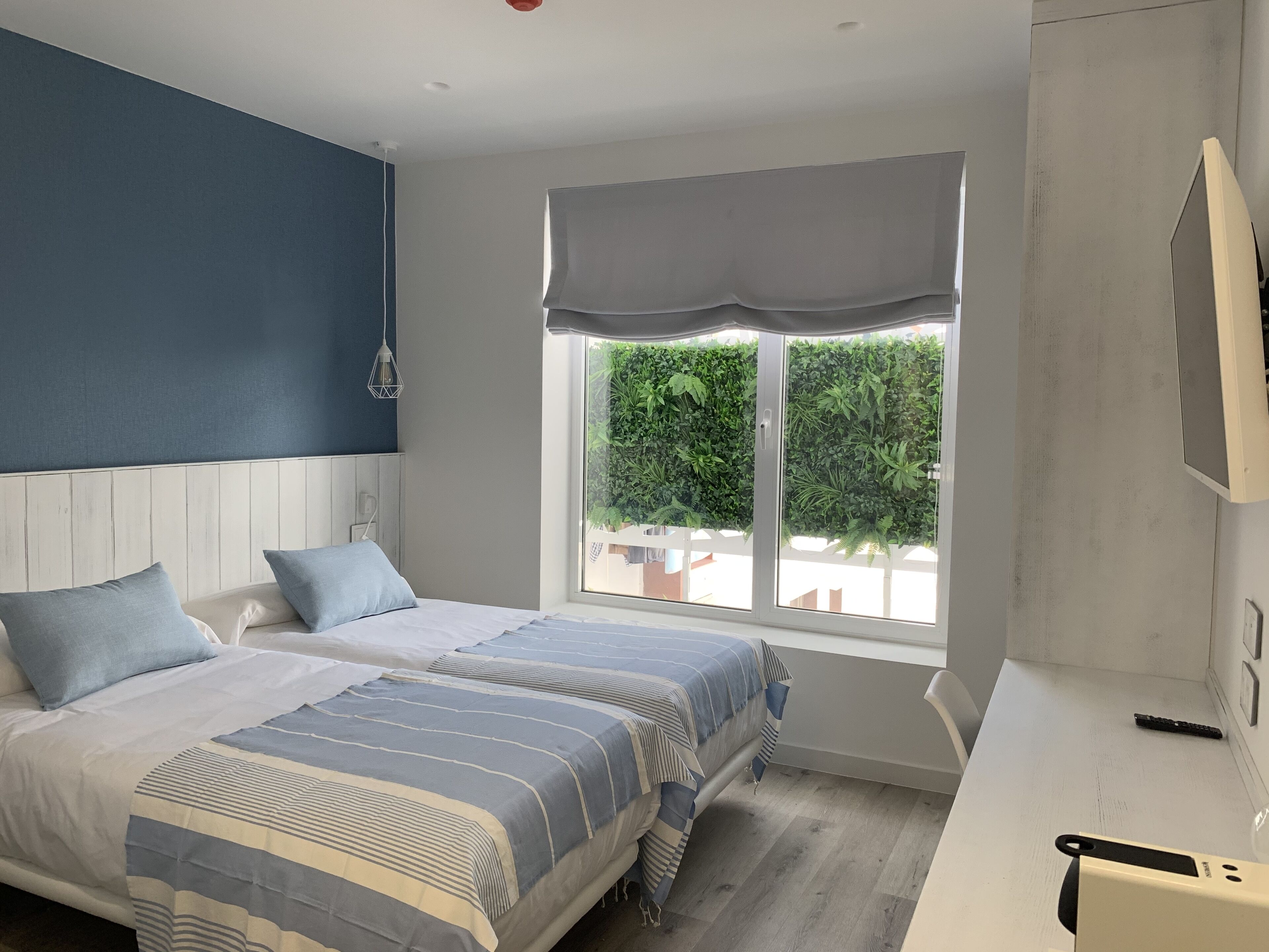 A bedroom with two beds, a window with a view of greenery, a white desk, and a TV on the wall.