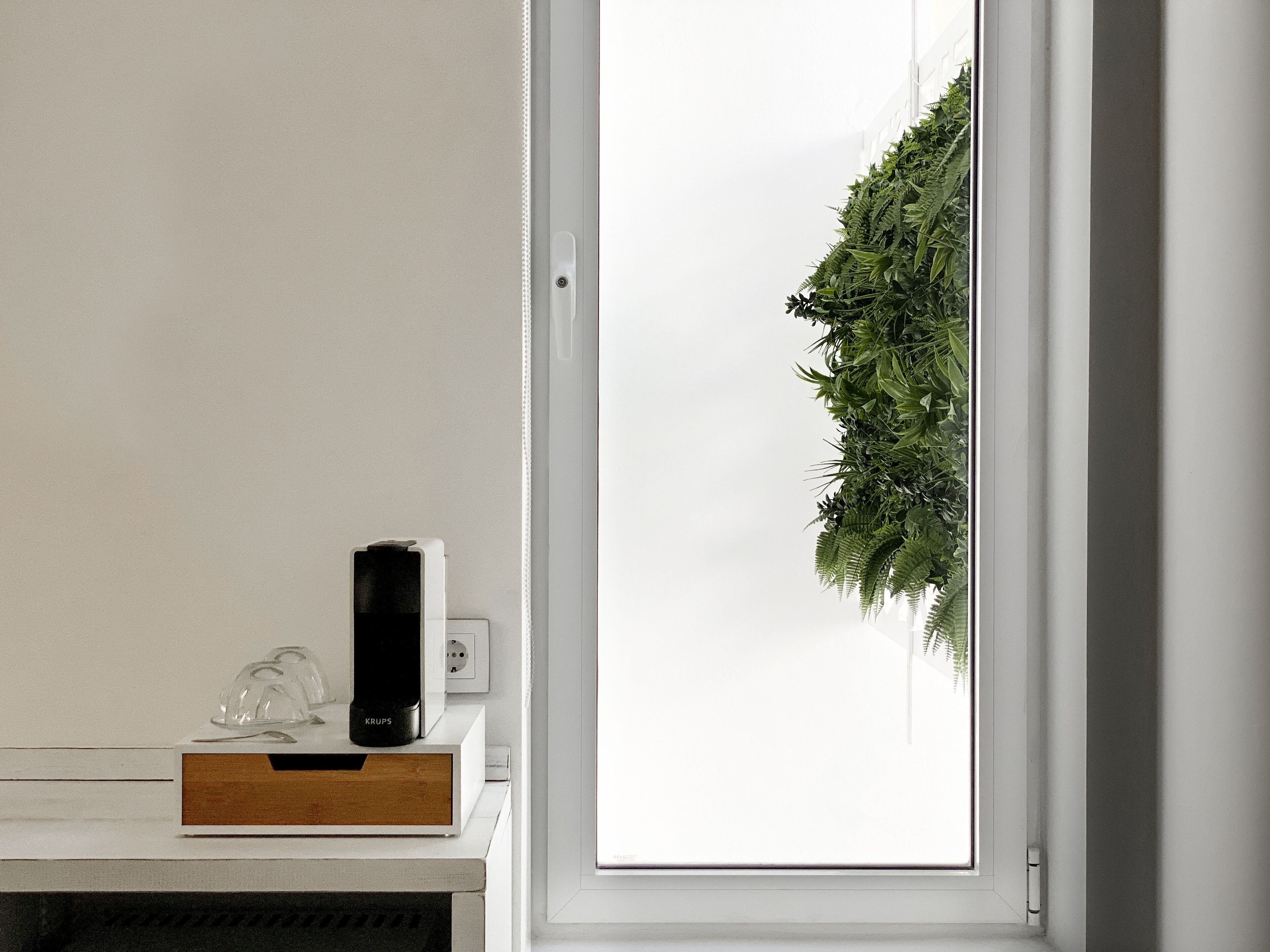 Une pièce avec une porte blanche, une petite table sur laquelle se trouve un objet noir, et une plante verte accrochée au mur.