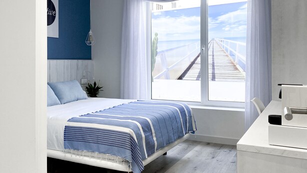 Une chambre à coucher avec un lit, une fenêtre donnant sur une jetée, une commode blanche et un mur orné d’un tableau encadré.