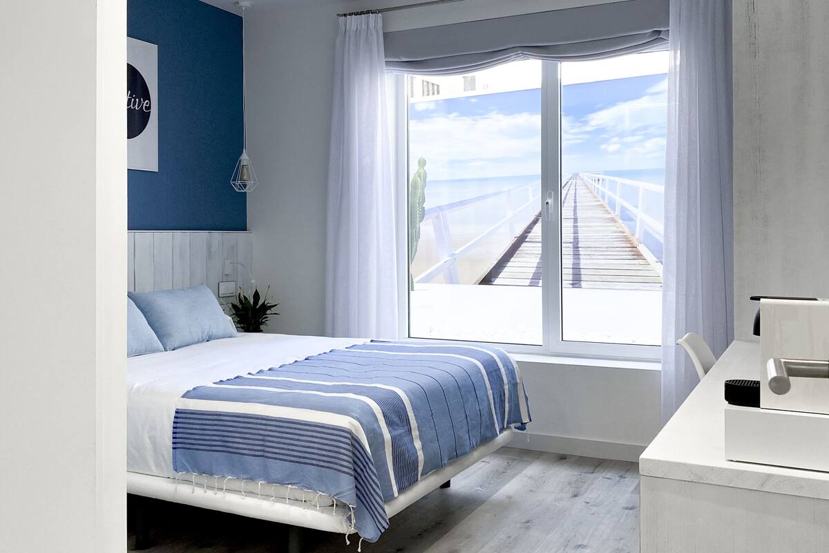 Un dormitorio con una cama, un ventanal con vista a un muelle, un tocador blanco y una pared con un cuadro enmarcado.