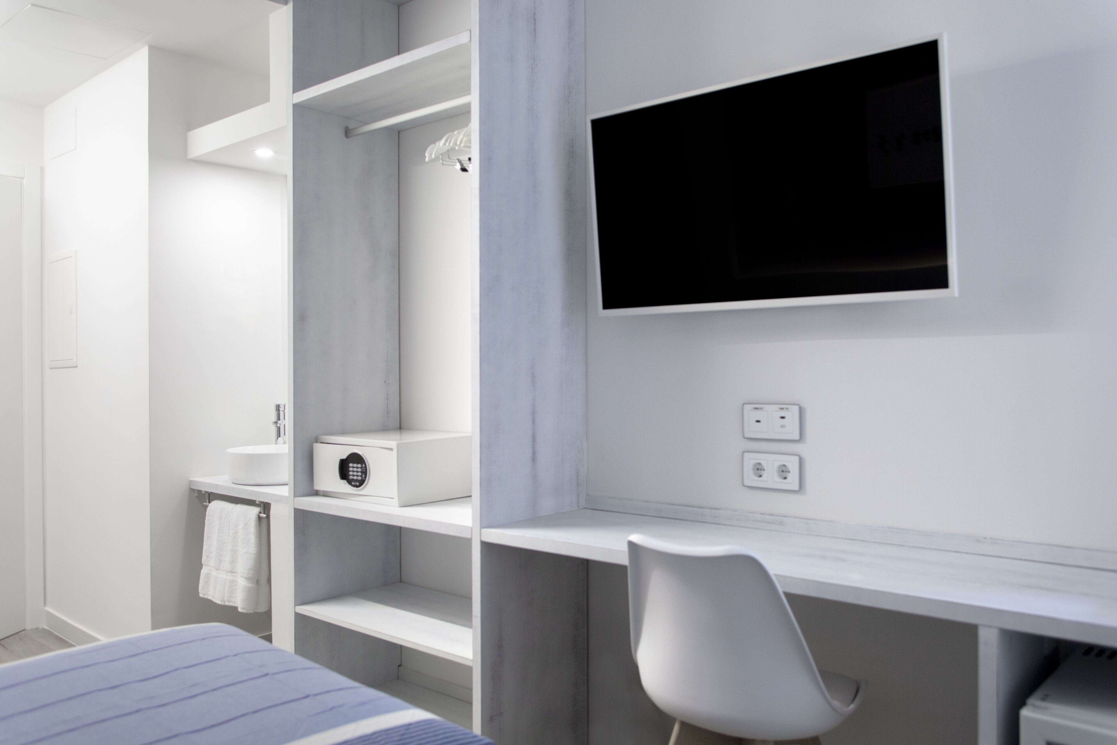 Quarto moderno com cama, escrivaninha, cadeira, televisão e banheiro com pia e toalha.