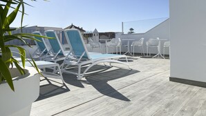 Une terrasse sur le toit, avec des chaises longues et des tables blanches, une plateforme en bois et un ciel d’un bleu limpide.