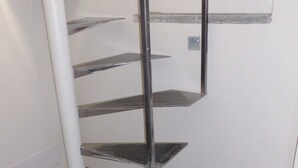 Een hoek van een kamer met een houten vloer, een witte muur en een metalen trap zonder treden.