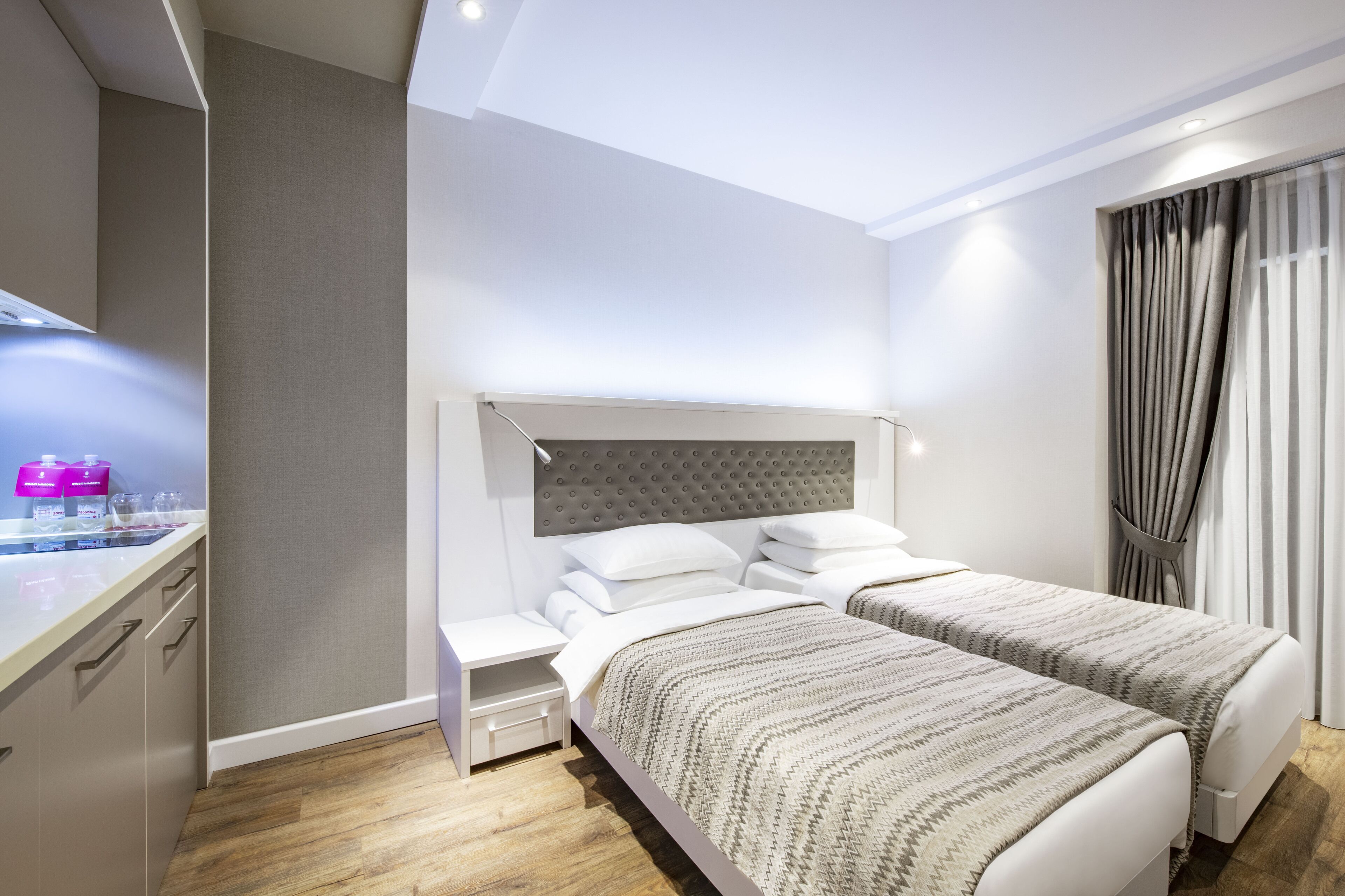 Quarto de hotel moderno com duas camas, uma mesinha de cabeceira, escrivaninha embutida e uma janela grande com cortinas.