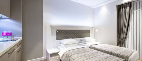 A modern hotel room with two beds, a bedside table, a built-in desk, and a large window with curtains.