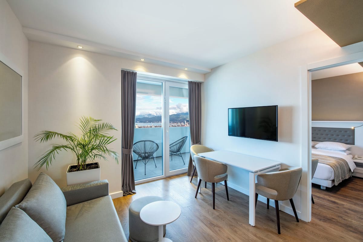 A modern hotel room with a balcony, a flat-screen TV, a sofa, a dining table, and a bed.