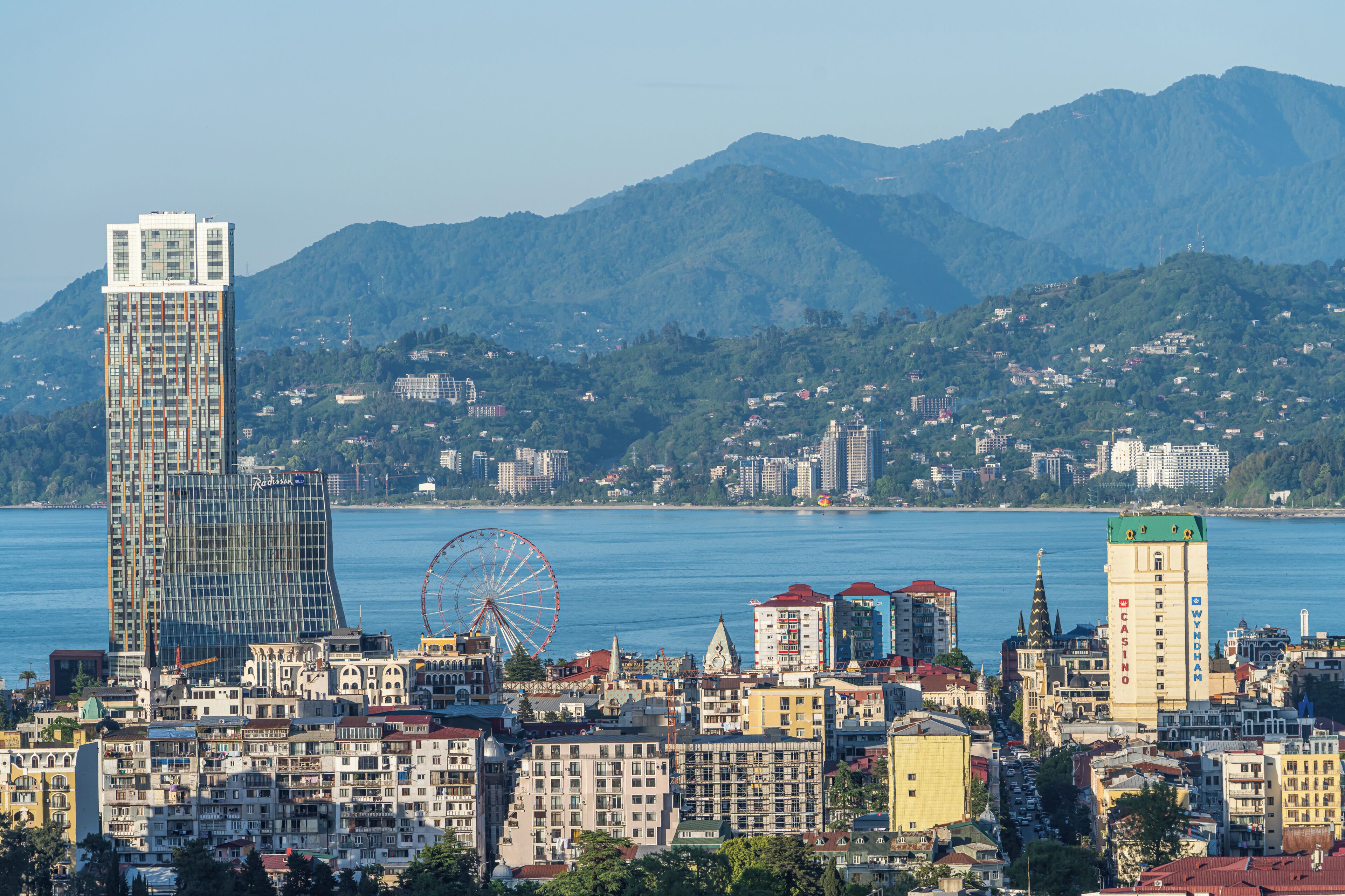 A cityscape with a prominent modern skyscraper, a Ferris wheel, and a body of water with mountains in the background.