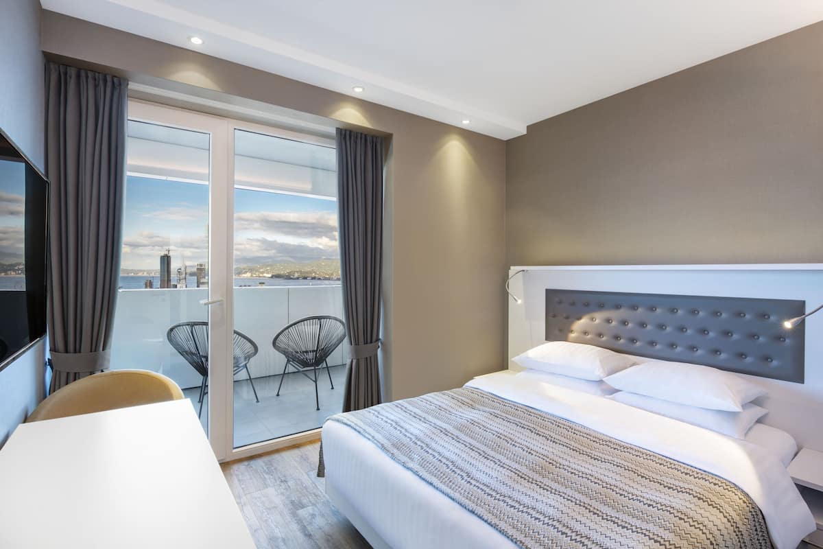 A hotel room with a large bed, a desk, and a view of the city through a glass door.